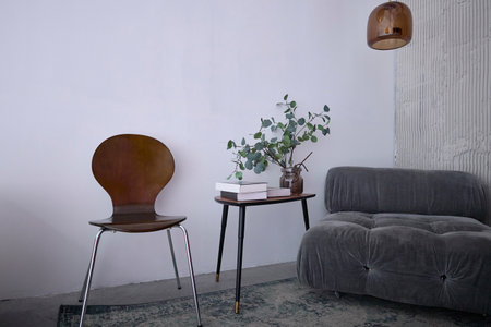 Modern minimalist living room featuring a gray sofa, wooden chair, and small table with books and a vase of greenery. Neutral tones and simple design create a relaxing atmosphere.の写真素材