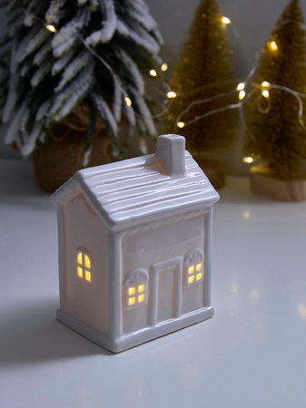 Ceramic house ornament emits a warm glow in a festive setting with string lights and snow-covered trees in the background, creating a cozy holiday ambiance.の写真素材
