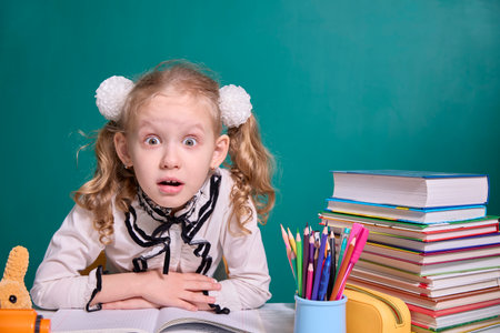 School girl with wide eyes and surprised expression surrounded by stacked books and colorful pencils. The image captures the essence of learning and curiosity in a classroom setting.の写真素材