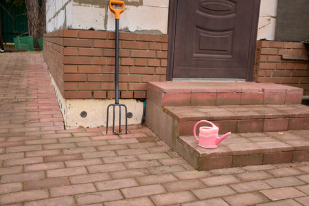 Brick patio with a garden fork leaning against the wall and a pink watering can on the steps. Outdoor setting, gardening scene, useful for illustrating home gardening topics.の写真素材