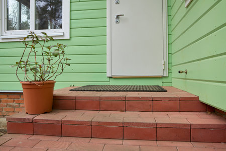 Green house exterior with a white door and steps made of red bricks. A potted plant sits on the steps, adding a touch of nature to the functional entrance area.の写真素材