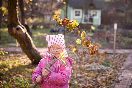 Smiling child in a pink jacket and hat playfully holds a rake with colorful autumn leaves in a garden setting, enjoying outdoor activities during the fall season.の写真素材
