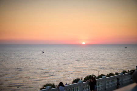 Two people on a balcony admire a serene ocean sunset, casting a warm glow on the tranquil sea. A boat sails in the distance, enhancing the peaceful atmosphere of the scene.の写真素材