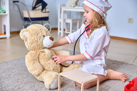 Child in doctor outfit playfully checks teddy bear's health using a stethoscope. Scene portrays imaginative play in a cozy room with toys, showcasing creativity and care.の写真素材
