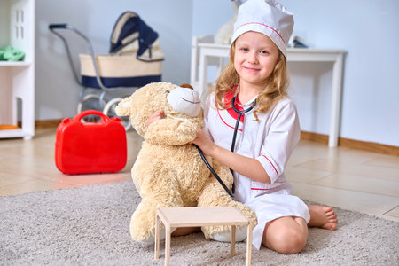 Young girl dressed as a doctor playfully checks a teddy bear's health with a stethoscope in a playroom setting, showcasing imaginative play and nurturing behavior.の写真素材