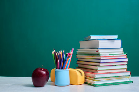 Stack of colorful books, a mug filled with colored pencils, an apple, and a yellow pencil case are arranged on a desk against a green chalkboard background, depicting academic theme.の写真素材