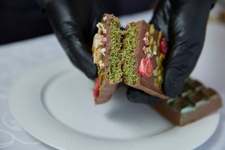Dubai chocolate. Delicate hands carefully break a textured milk chocolate bar, revealing pistachios and candied rose, a fleeting moment of handcrafted artistry, tempting indulgence.の写真素材