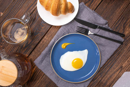 Fried eggs in a blue plate on a wooden table, breakfast with a croissant and freshly brewed tea. View from above.の写真素材