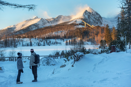 Unknown people standing near Strbske Pleso lake and looking far to the snowy mountain peaks of the High Tatras. Strbske pleso village is a favorite ski, tourist, and health slovakian resort.の写真素材