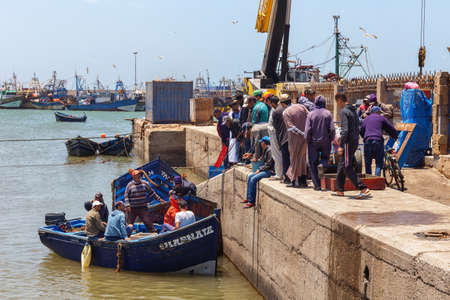 ESSAOUIRA, MOROCCO - JUNE 09, 2017: Unknown people on the pier in Essaouira harbor near the blue fishing wooden boat in the water.のeditorial素材