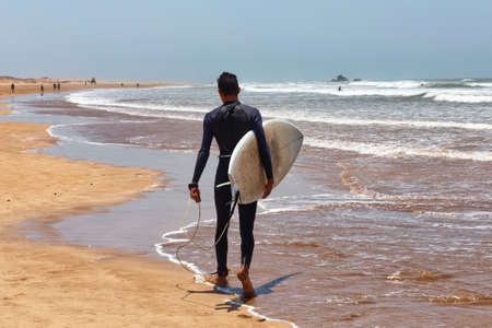 Unknown man in a wetsuit walking holding a surfboard in his hands along the Atlantic coast. Essaouira, Morocco.の写真素材