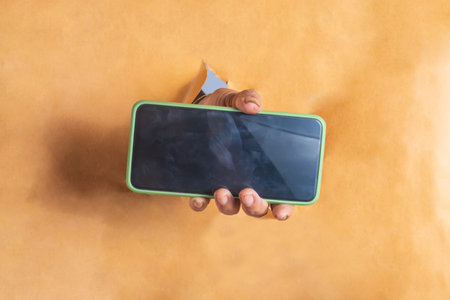 Close-up of woman hand holding mobile phone through torn paper wall. smartphone with blank screen