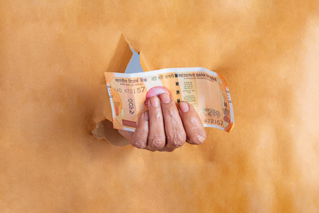 Hand of an elderly woman with a Indian 2oo rupee note coming out of torn brown paper.