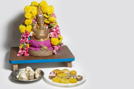 Ganesha statue made of clay decorated with flower garlands on wooden table and homemade sweets on white background. Copy space for textの写真素材