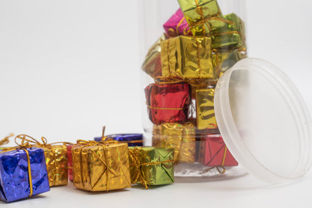 Colorful miniature gift boxes placed inside a clear plastic jar, lying on its side against a white...