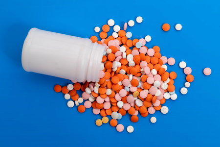 A variety of colorful pills and capsules spilled from an open container onto a clean, blue background. healthcare, medicine, and pharmaceutical concepts.の写真素材