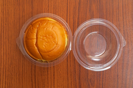 A single freshly made butter bun displayed inside a transparent plastic container on a wooden table. Perfect for presentations of fast food.の写真素材