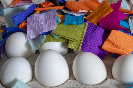 A carton containing twelve white eggs, surrounded by vibrant and colorful paper pieces.