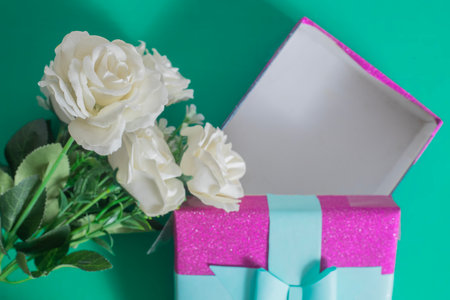 Beautifully arranged white roses next to a vibrant pink gift box decorated with a turquoise ribbon on a green background.の写真素材