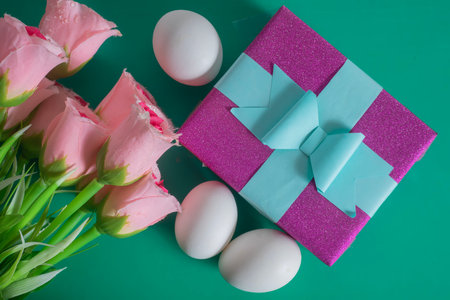 Top view of purple gift box with a blue ribbon, pink roses and white eggs on a green background.