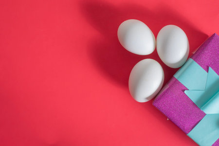 Three white eggs next to a vibrant, shiny, gift box with copy space for text, on a red surface.の写真素材