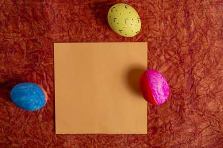 Brightly colored Easter eggs, placed on a rough orange surface around a plain orange paper. Evokes...