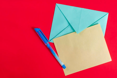 A blue envelope, a yellow piece of paper, and a blue pen artfully placed on a red background. Suitable for concepts related to communication, writing, and creativity.の写真素材