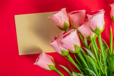 A bouquet of pink roses with water droplets arranged next to a plain gold card on a vibrant red background symbolizes love, celebration, and heartfelt messages.の写真素材