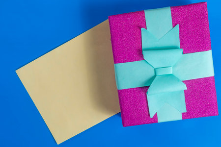 Pink glitter gift box with blue ribbon and blank card on vibrant blue surface for celebration, birthday, holiday or special occasions.の写真素材