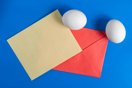 A vibrant arrangement with white eggs, a yellow paper sheet and a red envelope on a vibrant blue surface. Perfect for holiday, Easter or communication themes.の写真素材