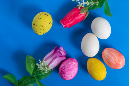 Brightly colored Easter eggs and artificial flowers are creatively arranged on a vibrant blue background for the Easter holiday season,の写真素材