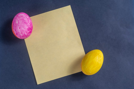 Brightly colored Easter eggs in pink and yellow next to plain yellow paper on a dark blue surface...
