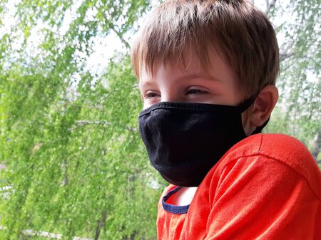 Kids in face mask sad near window. Boy in red T-shirt wear protective mask during coronavirus. Child stands on green summer foliage background.の写真素材