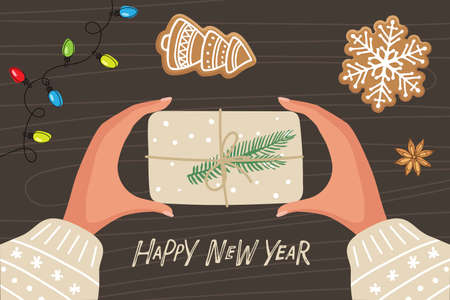 Two human hands holding box wrapped in kraft paper with decoration. Hands with Christmas gift, Christmas gingerbread, garland with lights on wooden table. Happy New Year lettering. Holiday decor.のイラスト素材