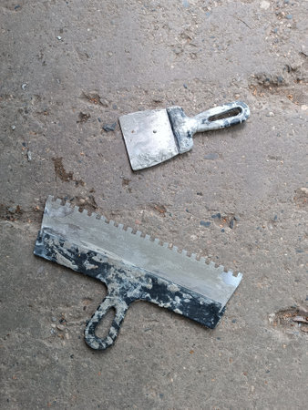 Two spatulas for laying tiles on concrete slab. Building tools lie on the floor. Dirty working tools. Wide spatula and small spatula with glue-stained handles on old potholed concrete. flooring conceptの写真素材