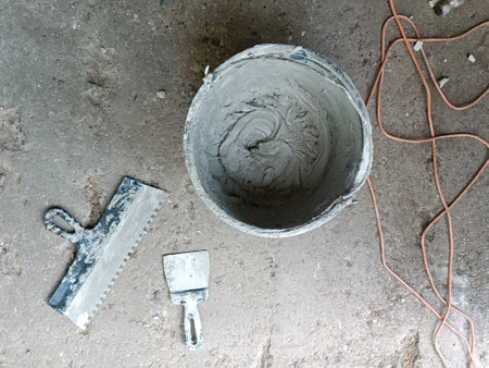 Black bucket with glue, spatulas on a concrete slab. Tools and materials for laying floor tiles. Old dirty black plastic bucket stands on floor. Renovation of old premises. Laying floor tilesの写真素材