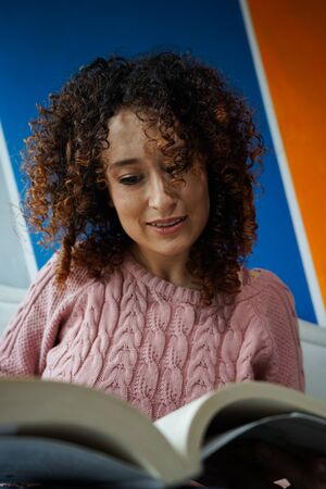 young woman in pink blouse and jean sitting on the armchair happily reading the pages of a book