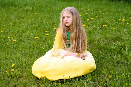 Blond young girl posing in a yellow green dress sitting on the grass with dandelions yellow flowersの写真素材
