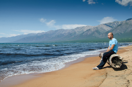 Men is sitting on a very old wooden rotten log aground on the beach, near lake Baikal, Russia, on a sunny day in the summerの写真素材