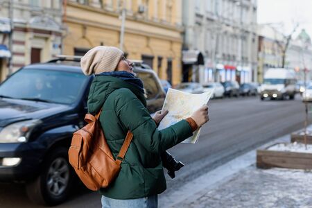 Girl in winter clothes with camera and cup of coffee in the historical center of the city. She is reading a map while visiting the city in the cold weatherの写真素材