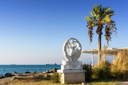 Turkey, Hatay - August 24, 2019: The Monument of white woman plays a pipe at iskenderun embankment with palm tree, sea, clear blue sky and ships in summer eveningのeditorial素材