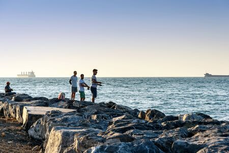 Iskenderun, Turkey - August 24, 2019: Sea fishing on spinning on the stone at seaside at sunset. Men casting lines on the Mediterranean coast with ship silhouetteのeditorial素材