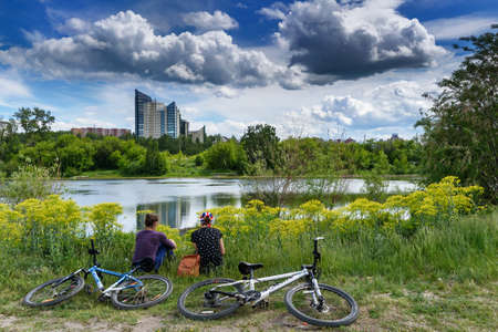 Two girls sit backs near their bikes. Background with modern building is reflected in lake in city Park. Summer Sunny day in a city with beautiful clouds. Irkutsk, Russia - June 8, 2020のeditorial素材