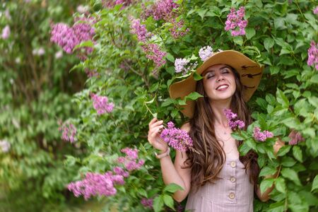 Girl in straw summer hat with closed eyes in pink dress enjoys summer against a background of blooming lilacの写真素材