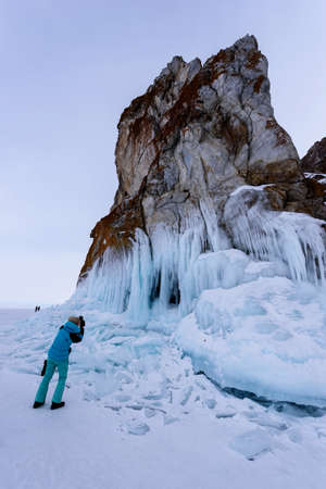 Lake Baikal, Russia - March 12, 2020: woman in a blue jacket takes photos of icy rocks with icicles on winter lake Baikalのeditorial素材