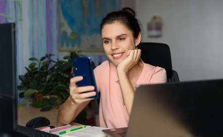 Smiling young woman working home near laptop, holds phone, smile saying sorry can not help with...
