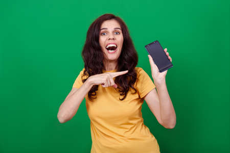 Young attractive woman is pointing on blank screen of phone in her hand and looks into a camera