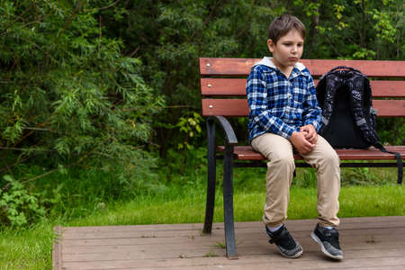 Sad school boy with backpack sitting on a street bench and thinkingの写真素材