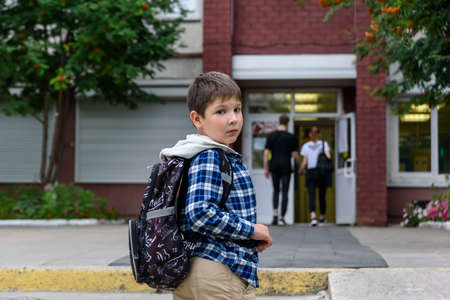 Child boy with bag stays near elementary school and looks to the camera