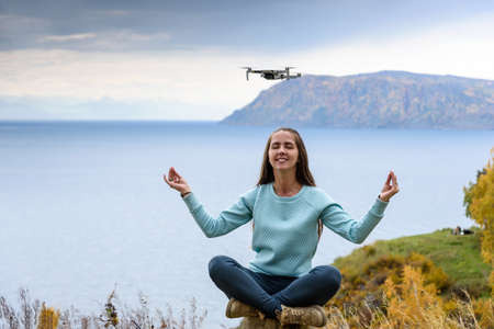 Beautiful young woman having fun with a mini drone outdoors in Lotus pose. The girl sits against the...
